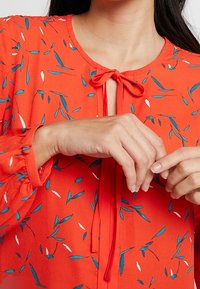 Röd blus med knyt vid halslinningen, med små blå och vita bladmönster, rynkade ärmavslut och en lätt, slät tygkvalitet.