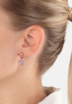 Nahaufnahme eines Ohres einer Frau mit einem silbernen herzförmigen Ohrring, der zwei klare Edelsteine enthält. Gepflegte blonde Haare nach hinten gebunden.