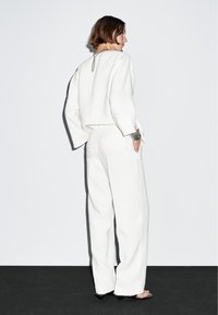 Vrouw die zijwaarts staat, gekleed in losse witte broek, witte blouse met lange mouwen, zwarte open hakken en een metalen armband om haar linkerpols.