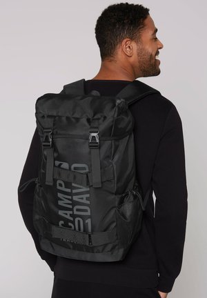 Black backpack featuring a roll-top design, adjustable straps, mesh side pockets, and a bold graphic logo on the front. Durable fabric texture.