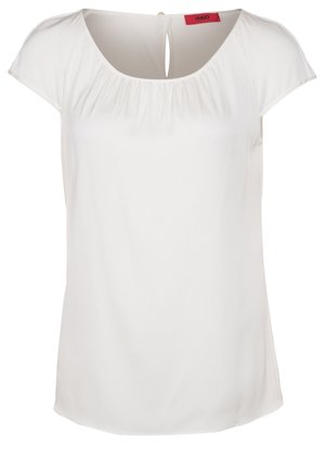 Blouse blanche à manches courtes avec encolure froncée et fermeture à bouton goutte d'eau dans le dos, avec une étiquette de marque rouge à l'intérieur du col.