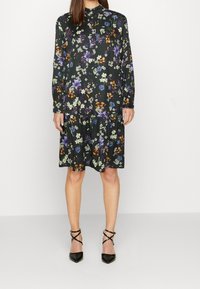 Nainen, joka on pukeutunut polvipituisen mustan kukkakuosisen mekkoon, jossa on pitkät hihat ja nappikaulus, yhdistettynä mustiin kärjellisiin korkokenkiin, joissa on nilkkaraidat.