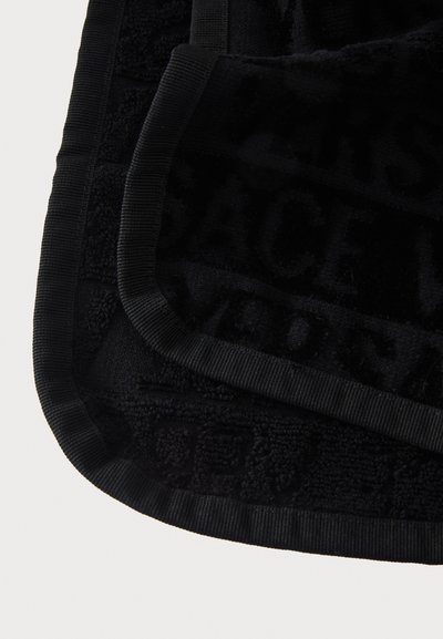 Detalii ale colțului unui prosop de baie negru pliat, cu text subtil în relief și margine rebarbată pe un fundal alb.