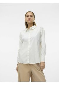 Camisa blanca de manga larga y botones con cuello, que presenta sutiles puntos texturizados, combinada con pantalones de pierna ancha en beige claro.