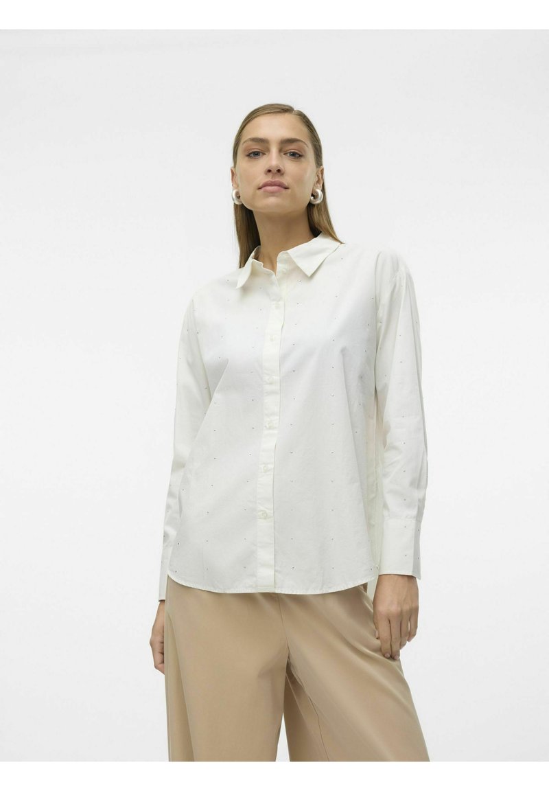 Camisa blanca de manga larga y botones con cuello, que presenta sutiles puntos texturizados, combinada con pantalones de pierna ancha en beige claro.