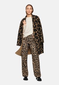 Leopardenmuster Mantel in Braun und Schwarz, kombiniert mit passenden Leopardenmuster Hosen und einem cremefarbenen Oberteil. Schwarze Schuhe runden das Outfit ab.
