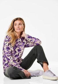 Langärmliges Oberteil mit einem geometrischen lila und beigen Muster. Kombiniert mit dunkelgrauen Jeans und helllila Sneakers. Glatte Stofftextur.