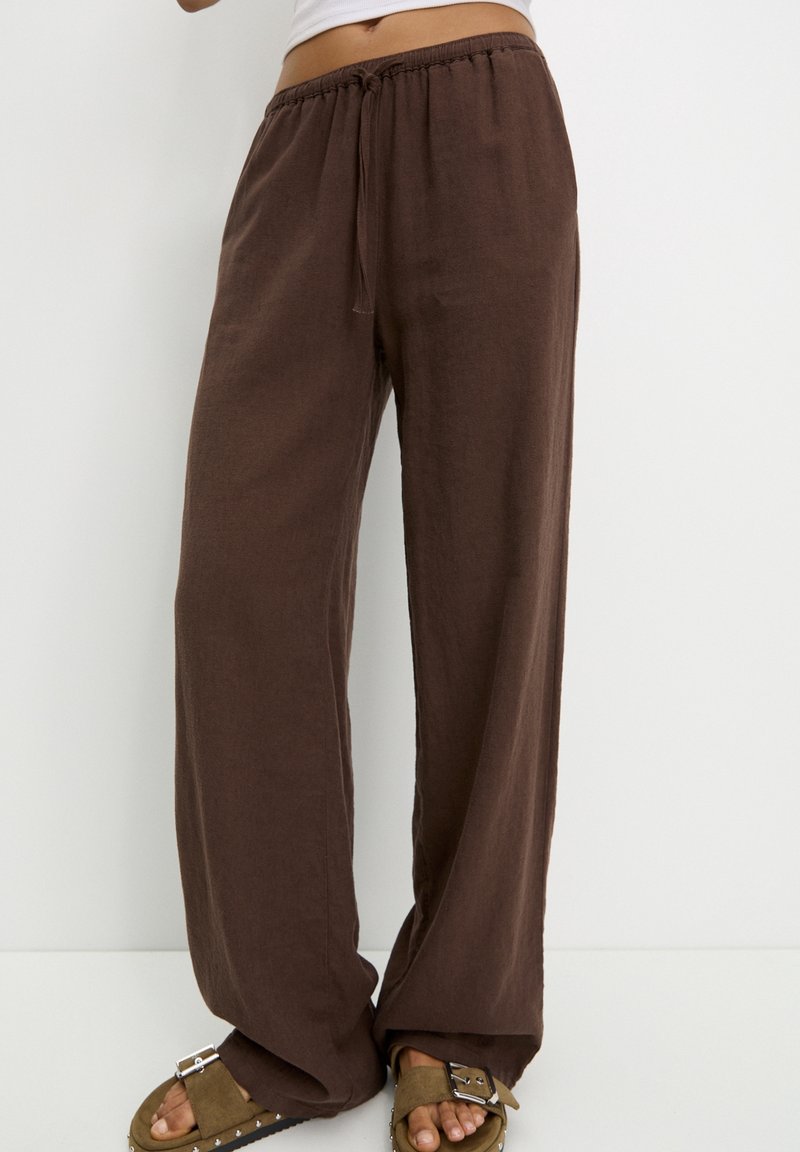 Personne portant un pantalon marron ample à cordon de serrage et des sandales vert olive avec des boucles argentées, debout sur un fond blanc uni.
