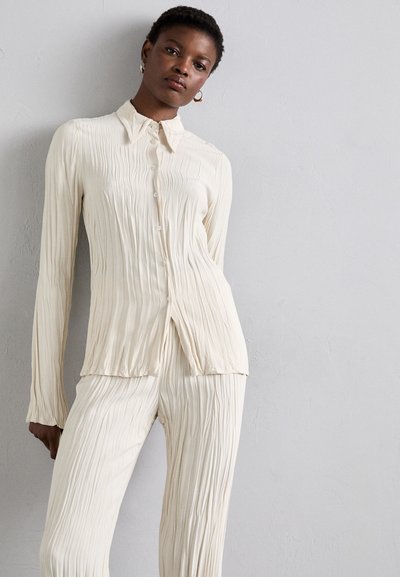 Cream-colored, pleated blouse with a button-up front, collar, and long sleeves, paired with matching pleated pants. Smooth texture.