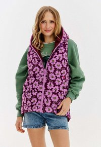 Młoda kobieta ubrana w purpurową, kwiatową kamizelkę wypełnioną powietrzem, pod którą ma zieloną bluzę i dżinsowe szorty, stojąca na białym tle.