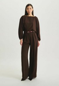 Combinaison marron à coupe décontractée, avec des manches larges et un décolleté carré, cintrée à la taille par une ceinture bordeaux. Accentuée par des jambes fuselées.