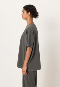 Mlada ženska z kodrastimi lasmi v figi, oblečena v ohlapno sivo majico s kratkimi rokavi in ujemajoče hlače, obrnjena levo na enobarvnem ozadju.