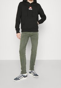 Sudadera negra con un pequeño logo blanco, pantalones verdes ajustados y zapatillas azules con rayas blancas. Outfit casual con un ajuste cómodo.