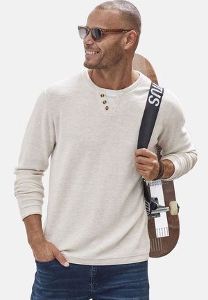 Hellbeige Langarm-Henley-Shirt mit drei Holzknöpfen, kombiniert mit dunkelblauen Jeans. Das Model hält ein Skateboard mit einem schwarzen Gurt.