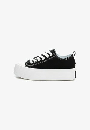 Zapatillas de lona negras con una gruesa suela de plataforma blanca, que presentan cordones blancos, detalles de costura y una lengüeta en la parte trasera.