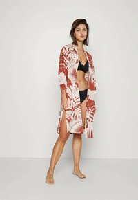 Woman standing barefoot wearing black bikini and knee-length open red and white floral patterned robe against plain background.