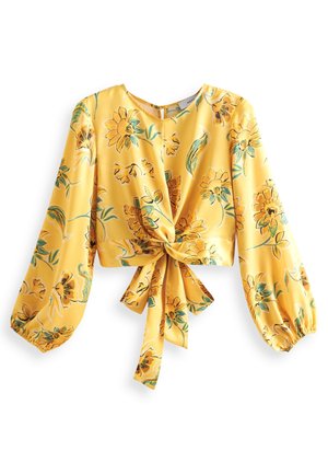 Blouse jaune à manches longues avec un nœud à l'avant, présentant de grands imprimés floraux orange et verts et des poignets élastiques.