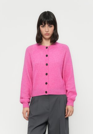 Femme portant un cardigan boutonné rose vif et un pantalon gris ample, se tenant devant un fond blanc uni.