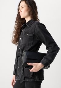 Črna usnjena torbica v roki, s strukturo, gladko teksturo in vtisnjenim logotipom 'ck', v kombinaciji s temno džins jakno.