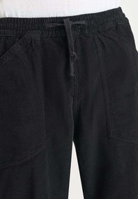 Zwarte corduroy broek met elastische tailleband en verstelbaar trekkoord, voorzien van grote voorzakken en zichtbare verticale ribbeltextuur.