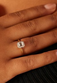 Gouden ring met een smaragdgeslepen centraal edelsteen omringd door kleinere stenen, rustend op een hand met een getinte, bruine huid.