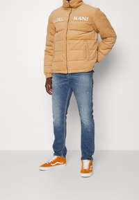 Persona che indossa un piumino beige con la scritta "KARL KANI", jeans blu e scarpe da skate arancioni brillanti, in piedi su uno sfondo bianco.