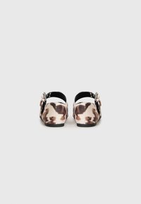Pair of flat shoes with brown and white cow-print pattern and black ankle straps with silver buckles, shown from the back.