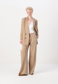 Lys brun skreddersydd blazer med svarte knapper, matchende vide benbukser, og en hvit v-hals topp, komplettert med en liten rosa håndveske.