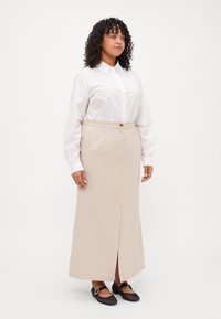 Lyst beige blyantkjole med splitt foran, med en midje og knappdetalj; kombinert med en hvit skjorte med krage og svarte sko.