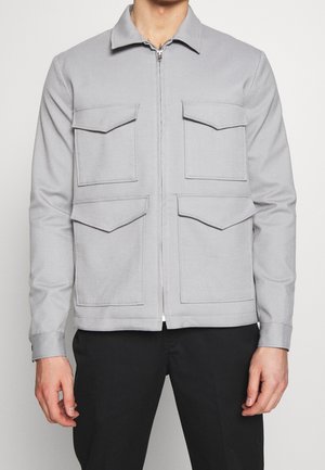 Uomo che indossa una giacca grigia chiaro con cerniera e quattro tasche con pattina sul davanti, pantaloni neri, su sfondo neutro.