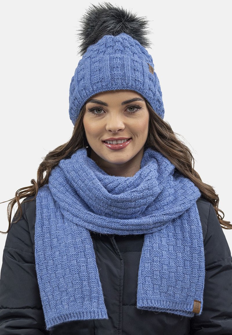 Căciulă tricotată albastră cu un pompon din blană sintetică și o eșarfă asortată. Ambele au un model texturat și o finisare moale.