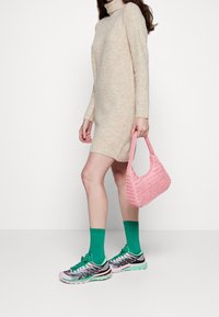 Robe en maille beige avec un col montant, associée à des chaussettes vertes et des baskets multicolores. Tenant un sac à main tissé rose.