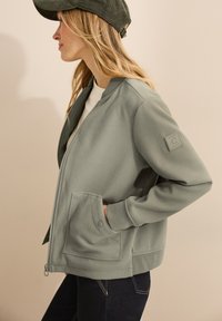 Veste bomber en tissu doux, de couleur verte, avec une fermeture éclair, des poches latérales et un écusson texturé sur la manche. Portée par-dessus une chemise blanche.