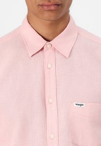 Šviesiai rožinė mygtukinė marškiniai, pagaminti iš tekstūruotos medžiagos, su klasikiniu apykakle, priekinėmis kišenėmis ir mažu Wrangler logotipu ant kišenės.