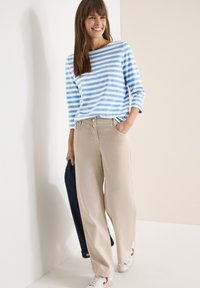 Femme souriante portant un haut à manches longues rayé bleu et blanc, un pantalon beige à jambes larges, des baskets blanches, tenant une veste en denim foncé à la main.