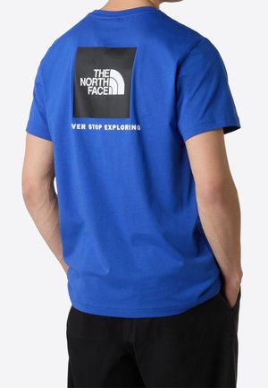 Man draagt een blauwe The North Face T-shirt met zwart logo en witte slogan "Never Stop Exploring" op de rug, gecombineerd met een zwarte broek.