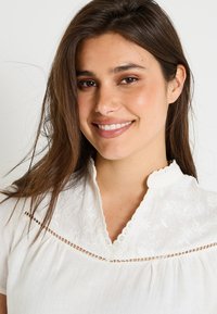 Blouse blanche avec un empiècement décoratif brodé, un col festonné et de fins plis, fabriquée dans un tissu léger et ornée de délicates accents en dentelle.