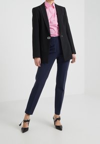 Blazer nero su misura con un'unica chiusura, camicia rosa con bottoni, pantaloni blu navy e décolleté neri a punta con dettagli in raso.