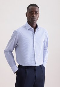 Camicia con bottoni, a righe verticali blu e bianche, maniche lunghe e colletto classico. Indossata con pantaloni sartoriali blu scuro.