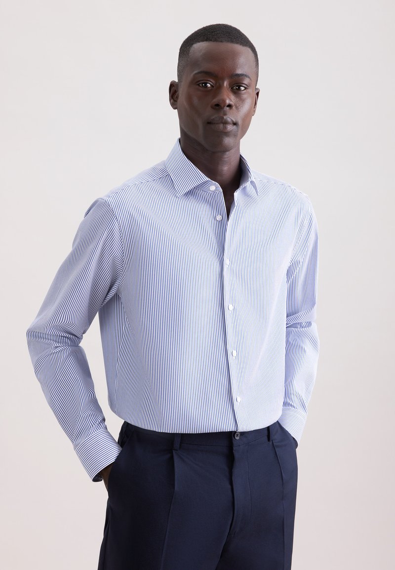 Camicia con bottoni, a righe verticali blu e bianche, maniche lunghe e colletto classico. Indossata con pantaloni sartoriali blu scuro.