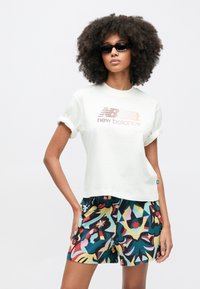 Biele bavlnené tričko s vyhrnutými rukávmi a ružovým logom "New Balance". Farebné vzorované šortky s abstraktnými tvarmi v modrej, červenej a žltej.