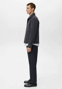 Giacca grigio scuro con colletto, tasche laterali e taglio dritto, indossata sopra una camicia azzurro chiaro e abbinata a pantaloni scuri e mocassini.