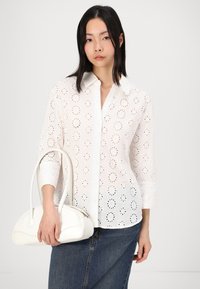 White cotton blouse featuring broderie anglaise design, pointed collar, and three-quarter sleeves. Model holds a white handbag with curved shape.