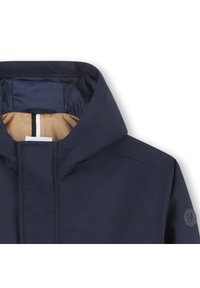 Marineblå jakke med hette, som viser en glatt tekstur, en kontrasterende tanfarget fôr og en rund logopatch på ermet.