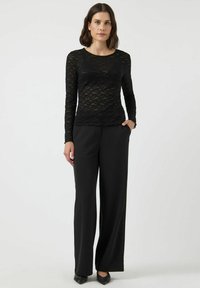 Svart långärmad spetsblus med ett texturerat blommönster, tillsammans med svarta vida byxor. Modellen står med händerna i fickorna.