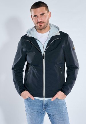 Man with short hair wearing layered black zip-up jacket over light gray hoodie and blue jeans, standing with hands in pockets.