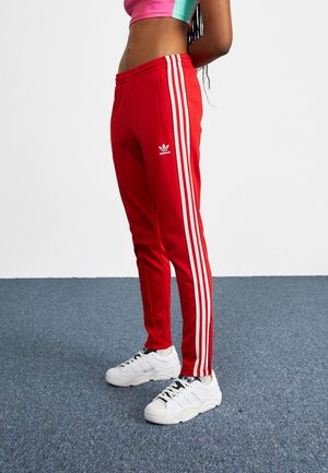 Personne portant un pantalon de survêtement rouge Adidas avec des bandes blanches et des baskets blanches, debout sur un sol recouvert d’un tapis bleu contre un mur uni.