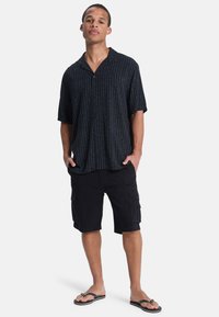 Krátký rukáv černé košile na knoflíky s modrými vertikálními pruhy, spárované s černými cargo šortkami a černými žabkami. Neformální letní outfit.