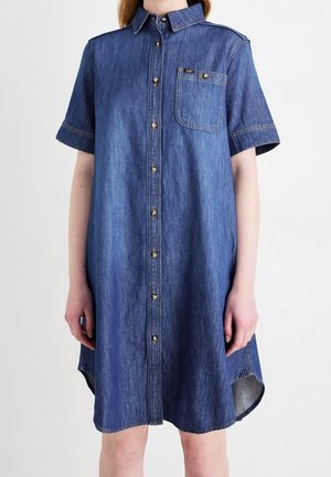 Modrá džínová košile s krátkými rukávy, předními knoflíky, náprsní kapsou a límečkem, nošená osobou stojící před jednobarevným pozadím.