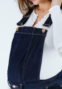 Donkere denim tuinpakken met gouden hardware, verstelbare banden en voorzakken, gedragen over een witte langemouwen top met een geknoopte halslijn.